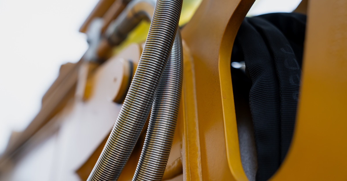 A close-up of an industrial macro of flexible metal hose and hydraulic line routed through a yellow excavator boom.