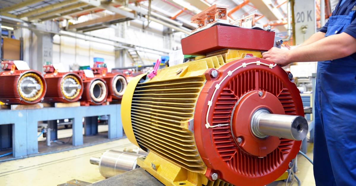 A person wearing blue work overalls stands beside a large red-and-yellow-colored electric motor mounted in a factory.