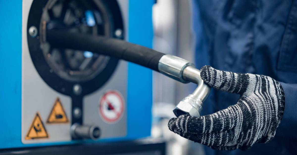 Someone wearing black and white gloves works with a hydraulic hose on a machine. The person holds a hose fitting in their hand.