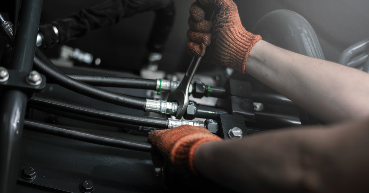 Someone wearing dirty orange work gloves is tightening a fitting around a hydraulic hose with a wrench.