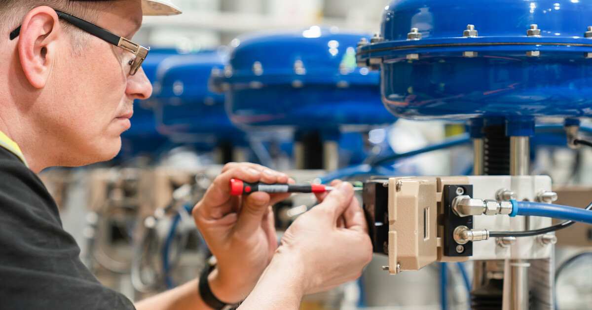 An individual in a white hard hat and a safety vest carefully holding a pneumatic valve while looking at it.