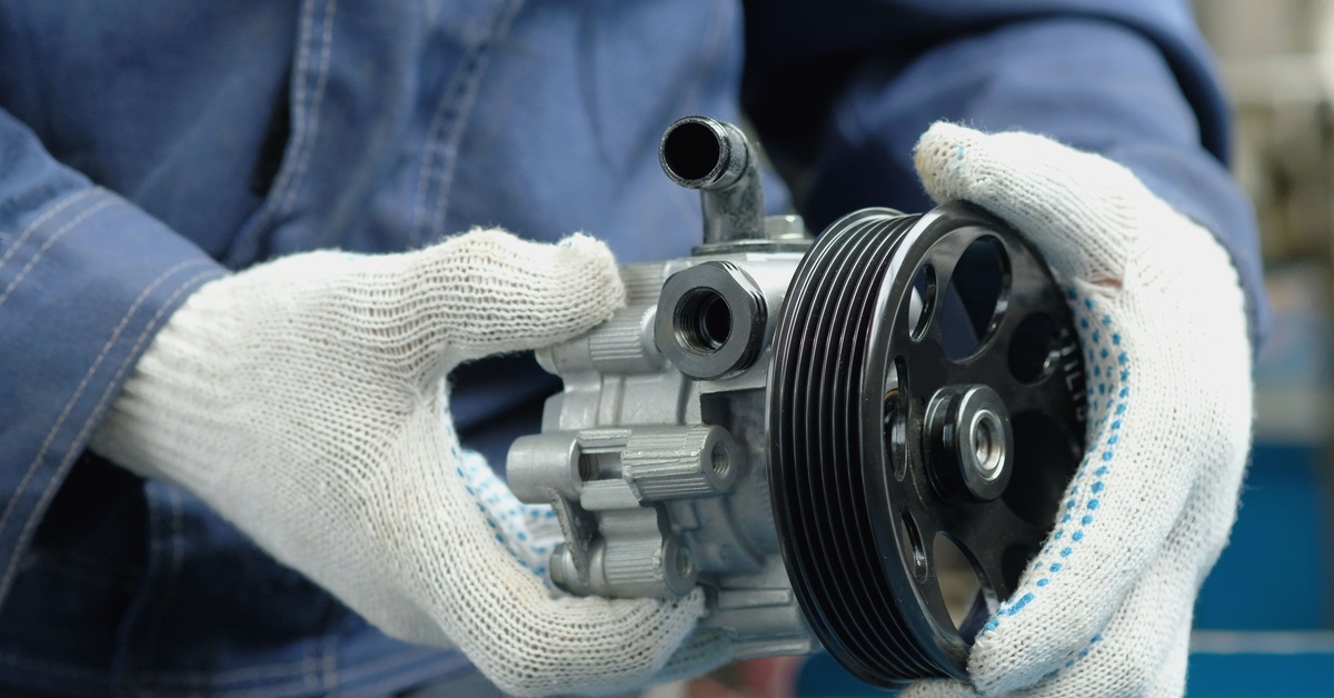 A person wears gray heavy-duty gloves as they hold a hydraulic steering pump in both their hands.