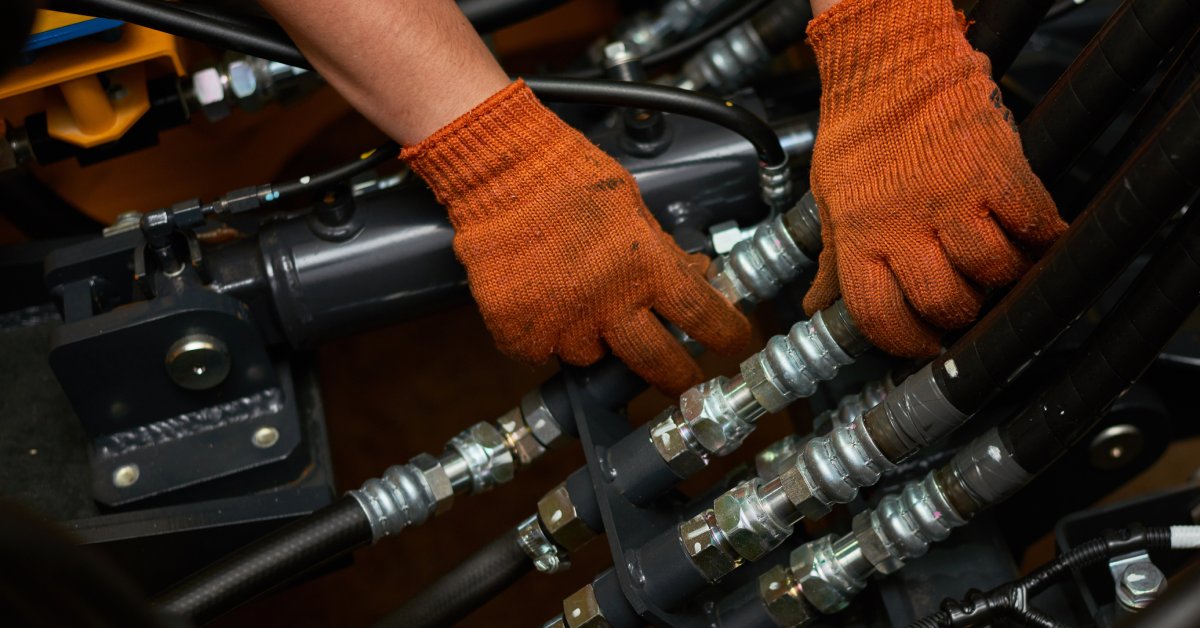 A close-up of a person wearing orange work gloves touching several industrial hydraulic hoses and pipes.