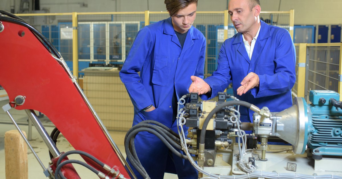 Two people in blue jumpsuits stand together and look at an industrial hydraulic arm mechanism with hoses and fittings.