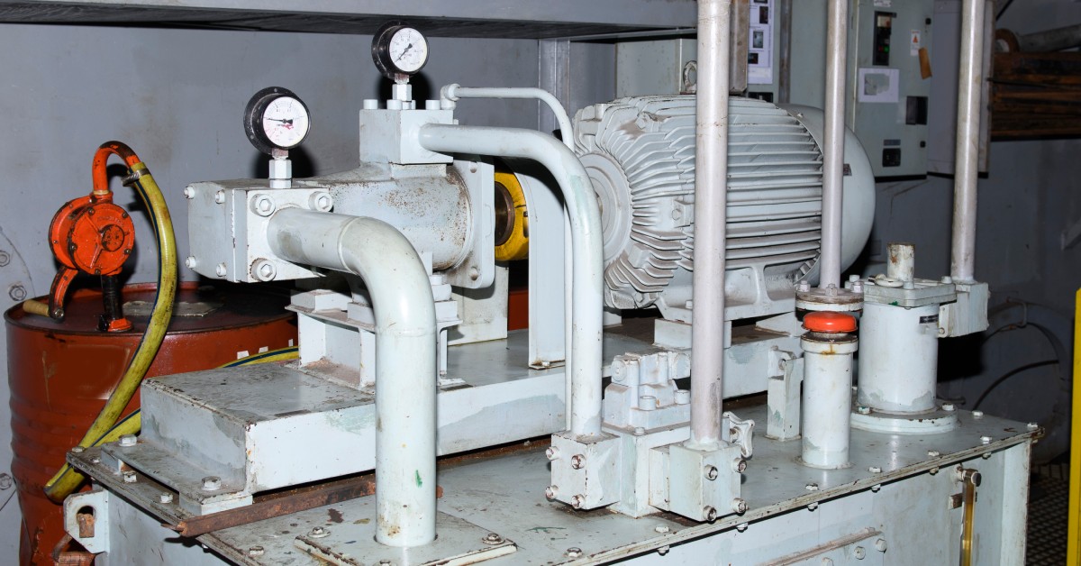 The steering room of a large vessel containing a slightly rusted-over hydraulic pump and rudder machine.