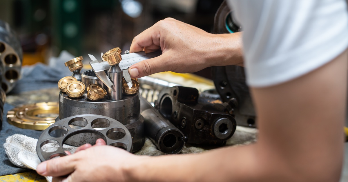 A person in a white T-shirt holds the piston of a hydraulic pump and examines machine parts laid out on a table.