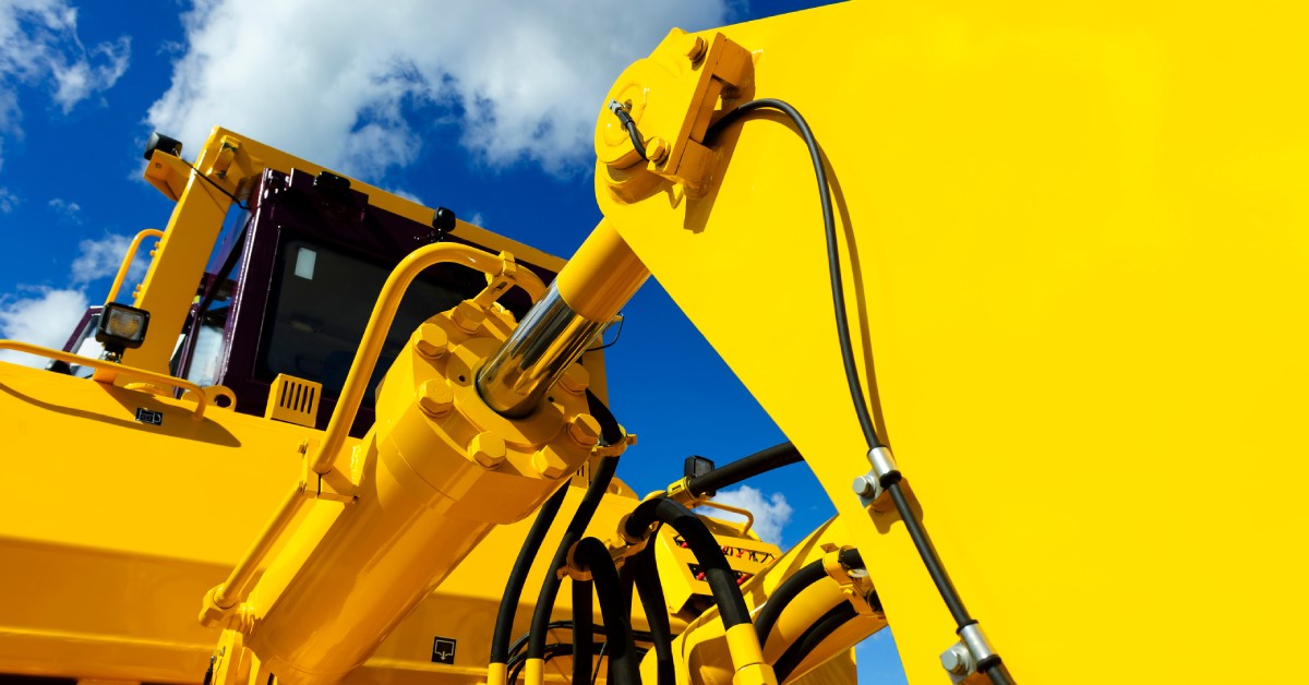 Close-up of a large yellow construction machine with a hydraulic piston arm against a blue sky with clouds.